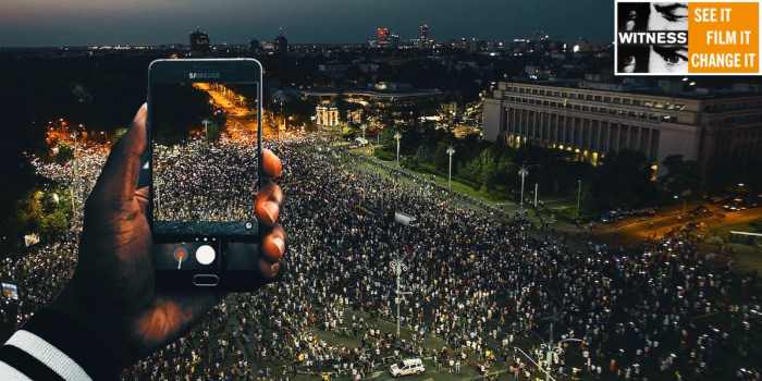 a man holds up a camera phone from a balcony to film protesters in the street at night time