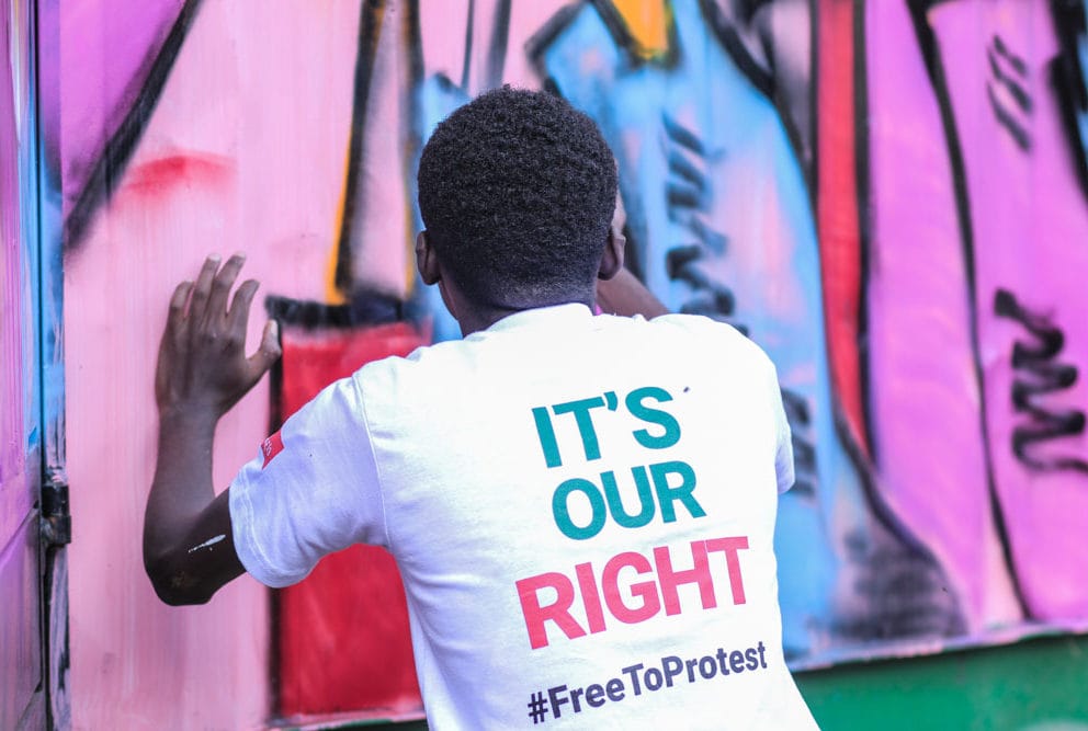 A grafitti artist paints a pink wall with a print on the back of his t shirt displaying the right to protest text