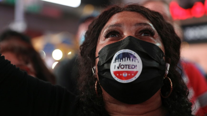 An African-American women wearing a mask with the word 'Voted, raises her face in celebration.