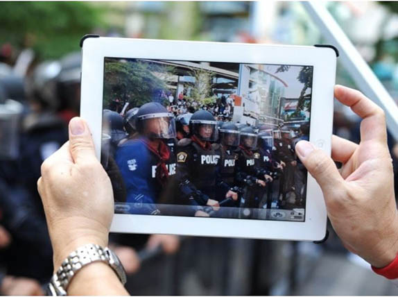 Hands holding a tablet which shows on the screen they are recording riot police who are standing in front 